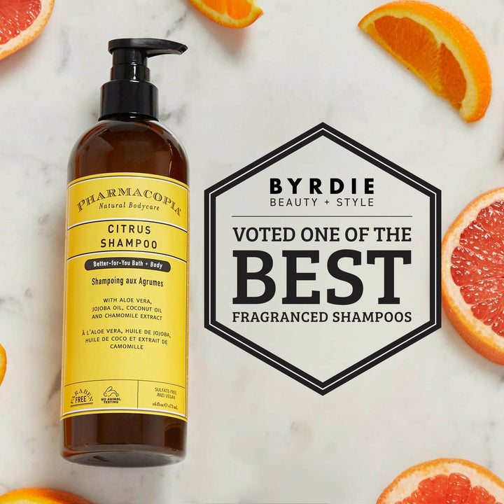 A bottle of Pharmacopia Citrus Shampoo stands on a light marble surface with orange and grapefruit slices around it. A large badge reads 'BYRDIE BEAUTY + STYLE VOTED ONE OF THE BEST FRAGRANCED SHAMPOOS.